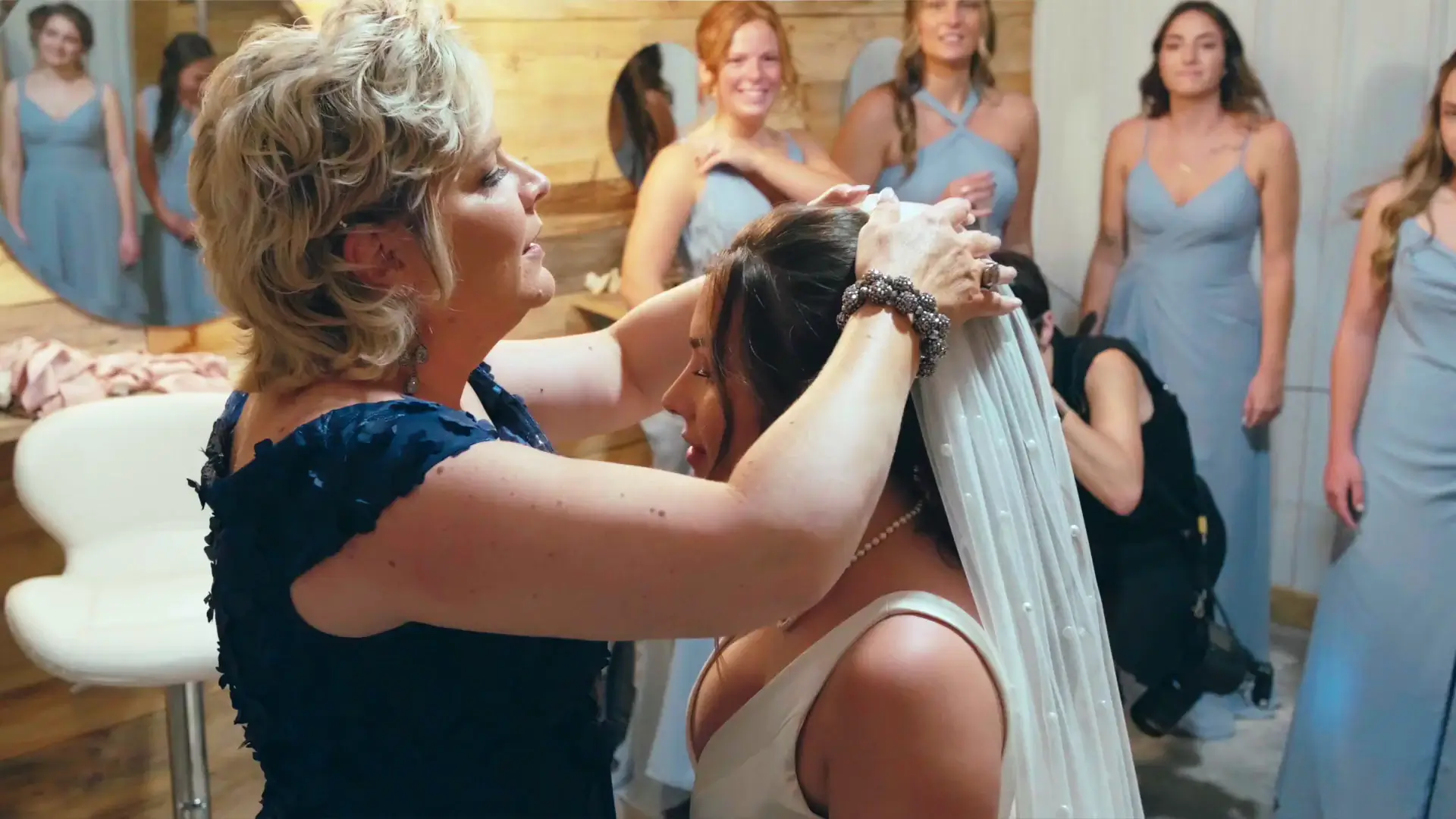 A woman adjusts the bride's veil surrounded by bridesmaids in light blue dresses. In this still from their wedding video, the intimate preparation moment is captured by their wedding videographer.