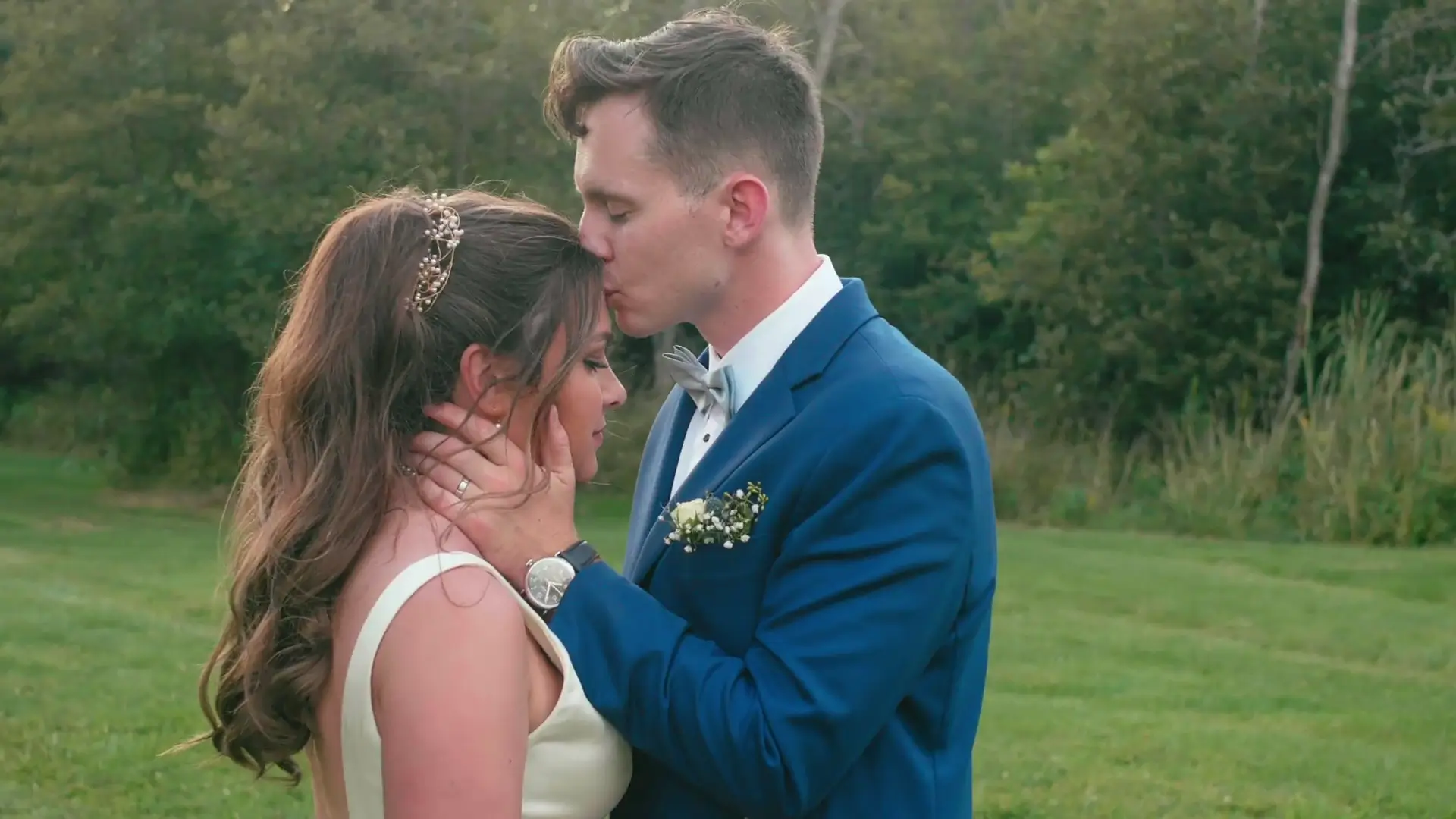In this still from their wedding video, a groom tenderly kisses the forehead of his bride as they stand on a grassy lawn, surrounded by greenery.