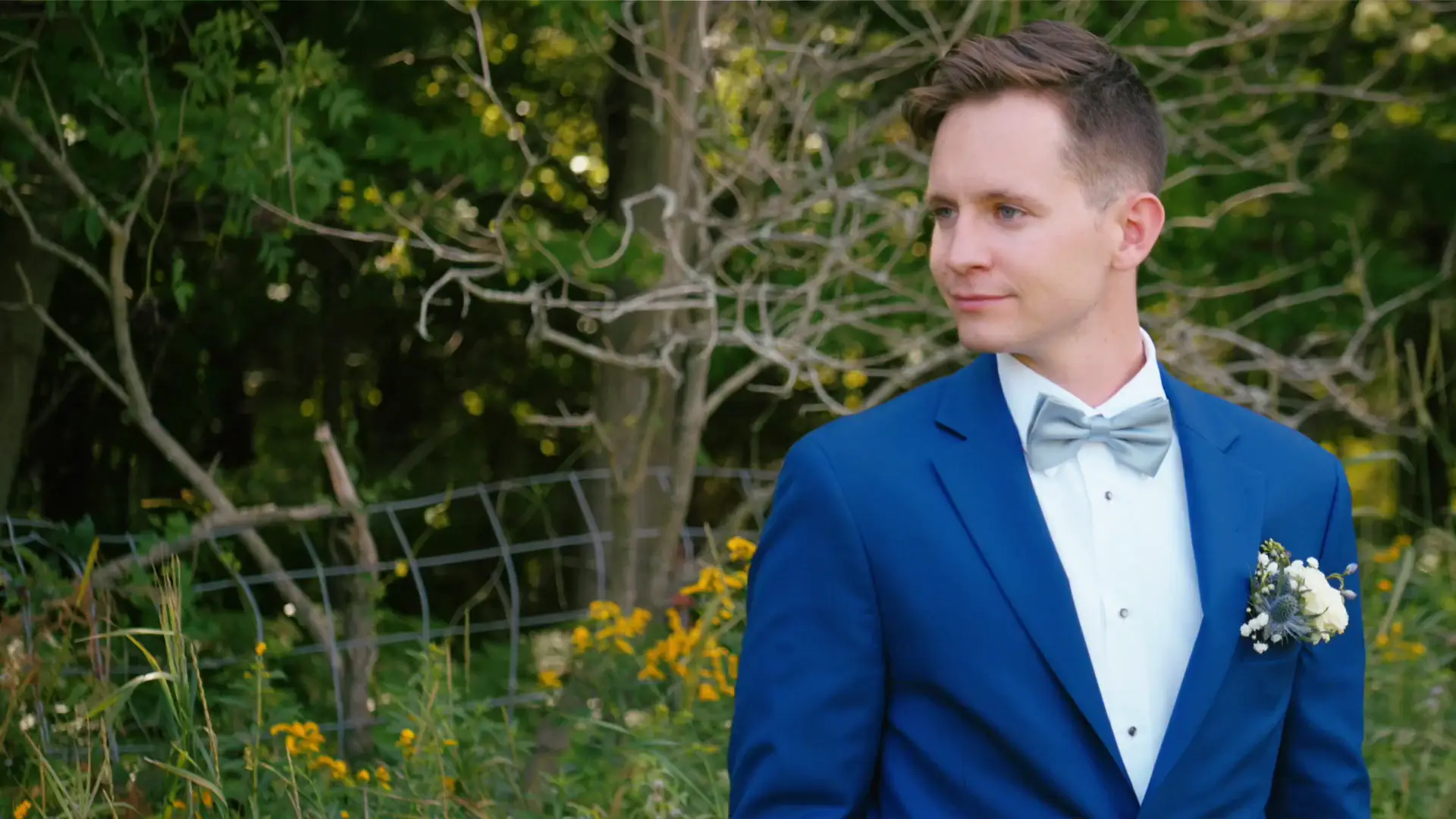 In this still from their wedding video, a person in a blue suit with a bow tie stands outdoors among green foliage and yellow flowers.