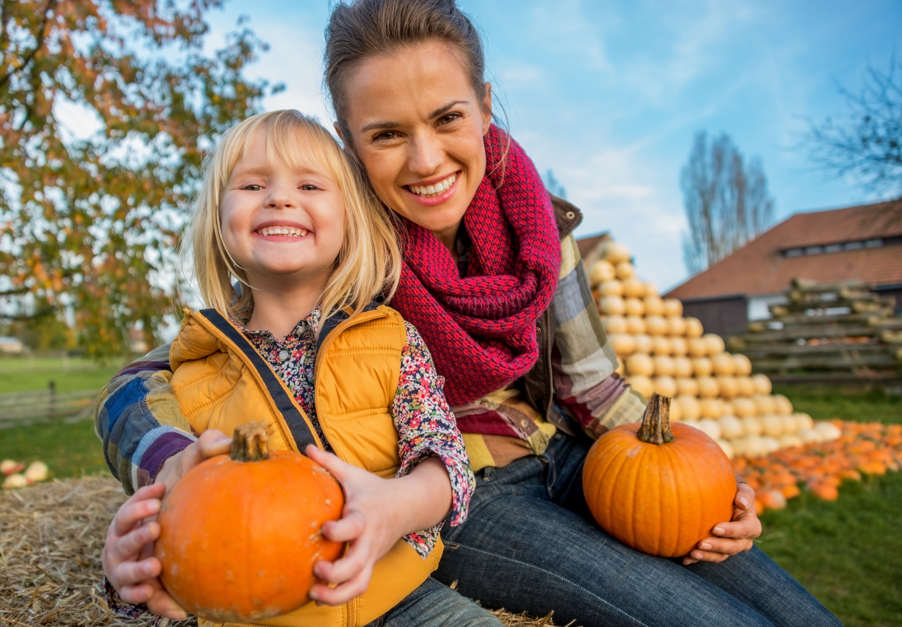 An adult and a child smiling while each holding a pumpkin on a hay bale