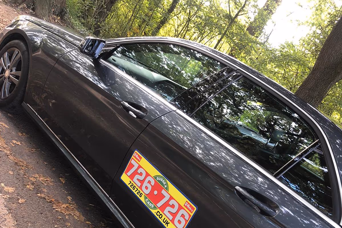 A parked black car with a yellow and red advertisement sticker on the door in a wooded area.