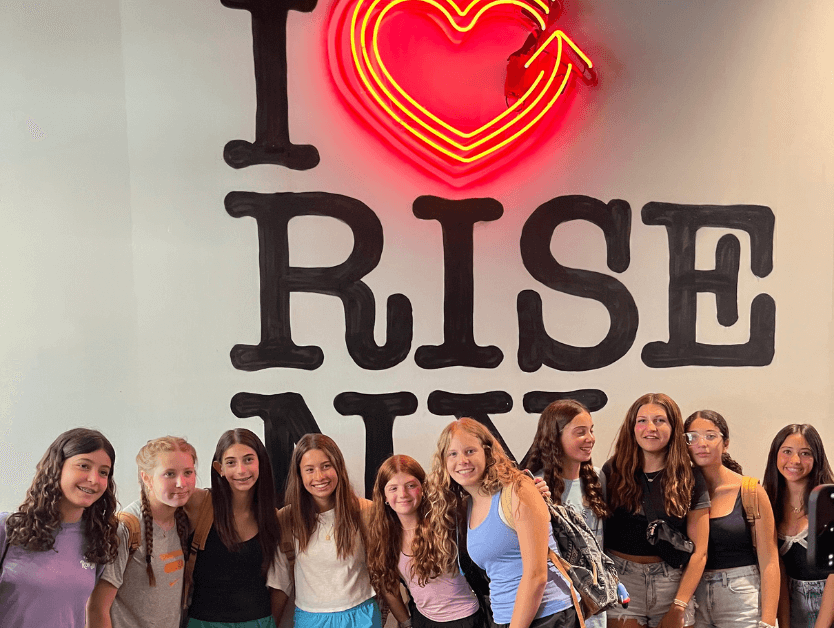 A group of students smiling and posing for a photo in front of the neon RiseNY sign
