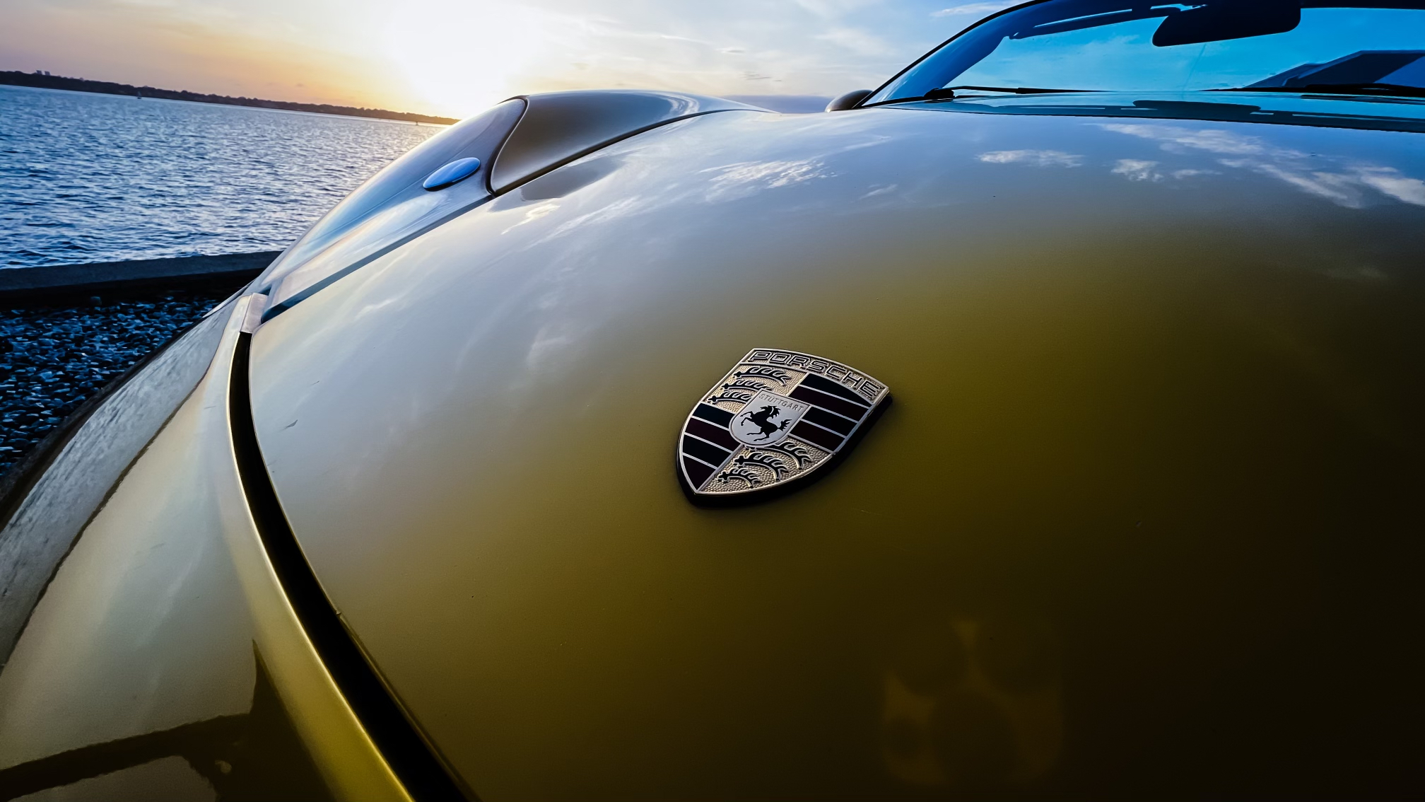 Close-up of Porsche emblem on the hood of a yellow sports car at sunset by the water in Newport RI, showcasing luxury car rental branding and coastal elegance.