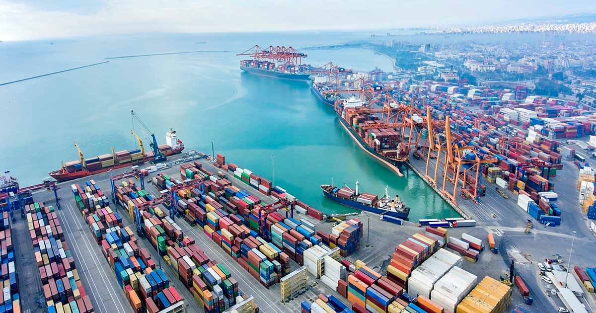 An overhead shot of a shipping port that features many shipping containers and five docked ships. | Sourcengine