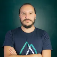 Man with beard and mustache wearing a dark blue t-shirt with a geometric logo on a dark green background.
