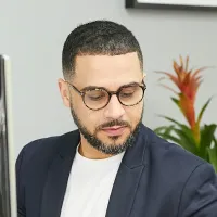 Man with glasses and beard wearing a dark blazer and white shirt, looking down.