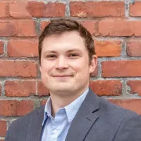 Smiling man in a gray suit jacket and light blue shirt standing in front of a brick wall.