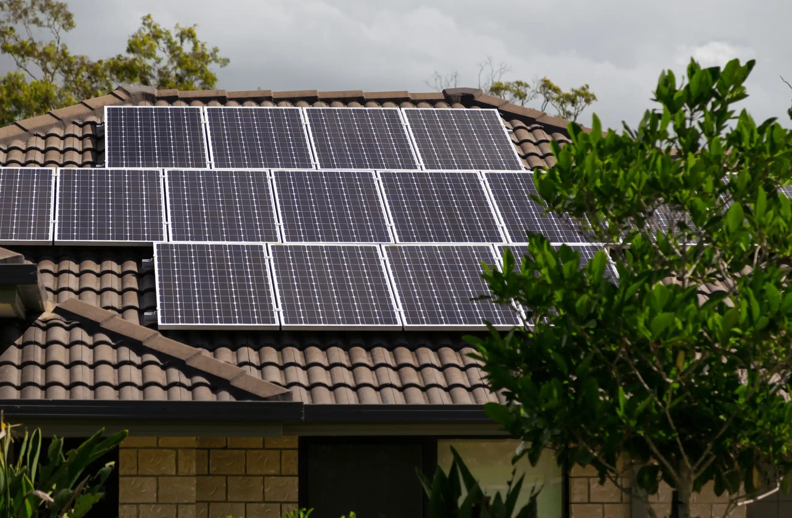 Solar panels are installed on the tiled roof of a house surrounded by green trees and plants, under a cloudy sky.