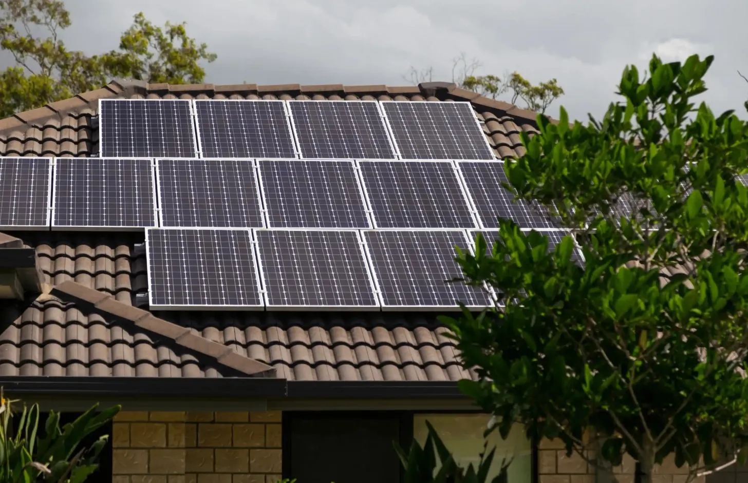 Solar panels are installed on the tiled roof of a house surrounded by green trees and plants, under a cloudy sky.