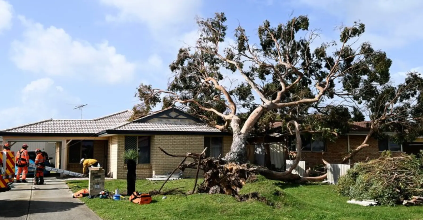 A large tree has fallen onto a suburban house, causing visible damage. Emergency workers in orange uniforms are assessing the scene, with tools and debris scattered on the lawn under a partly cloudy sky.