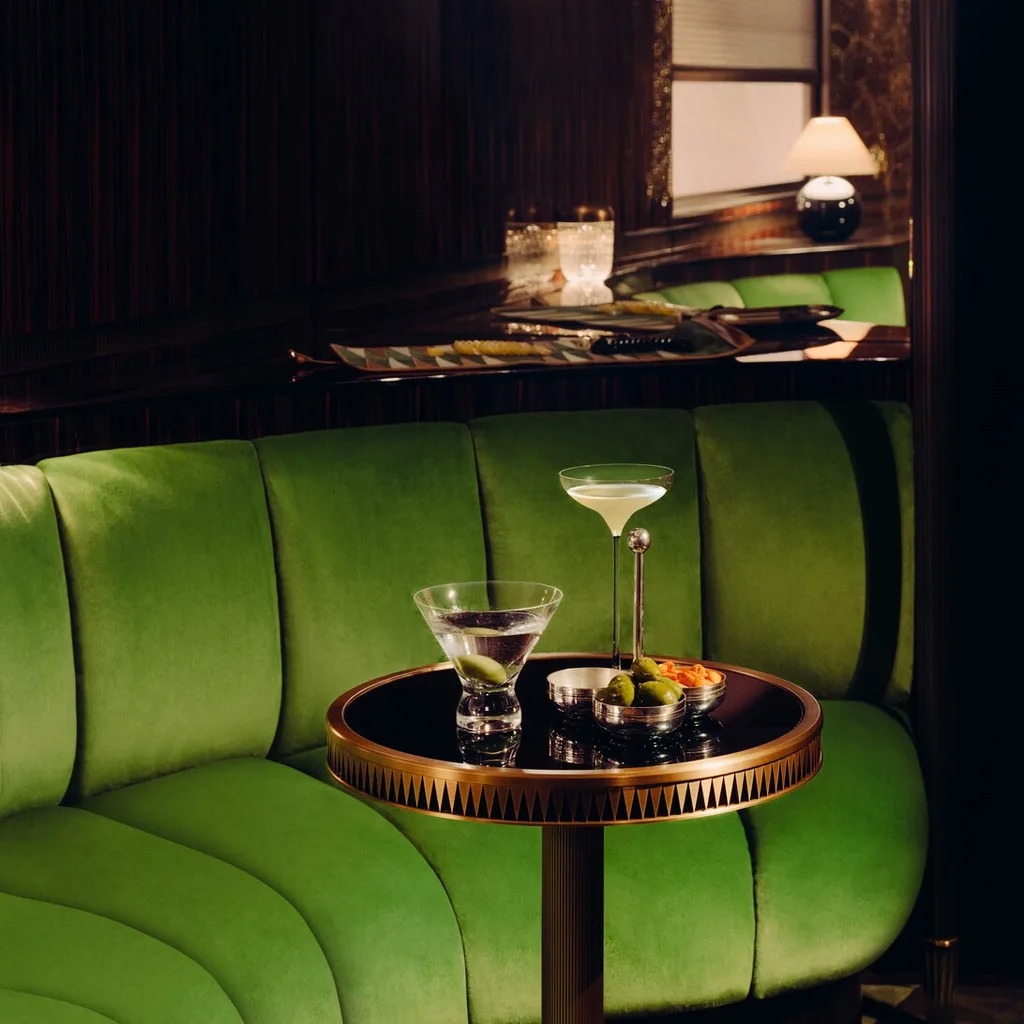 A small side table with olives and cocktails in front of velvet green banquettes.