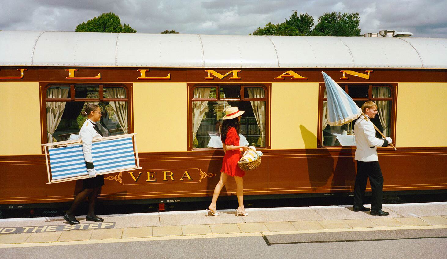 Staff for the British Pullman, holding an umbrella and sun-deck chair, leading and flaking a passenger holding a picnic basket on a train station platform outside the train.