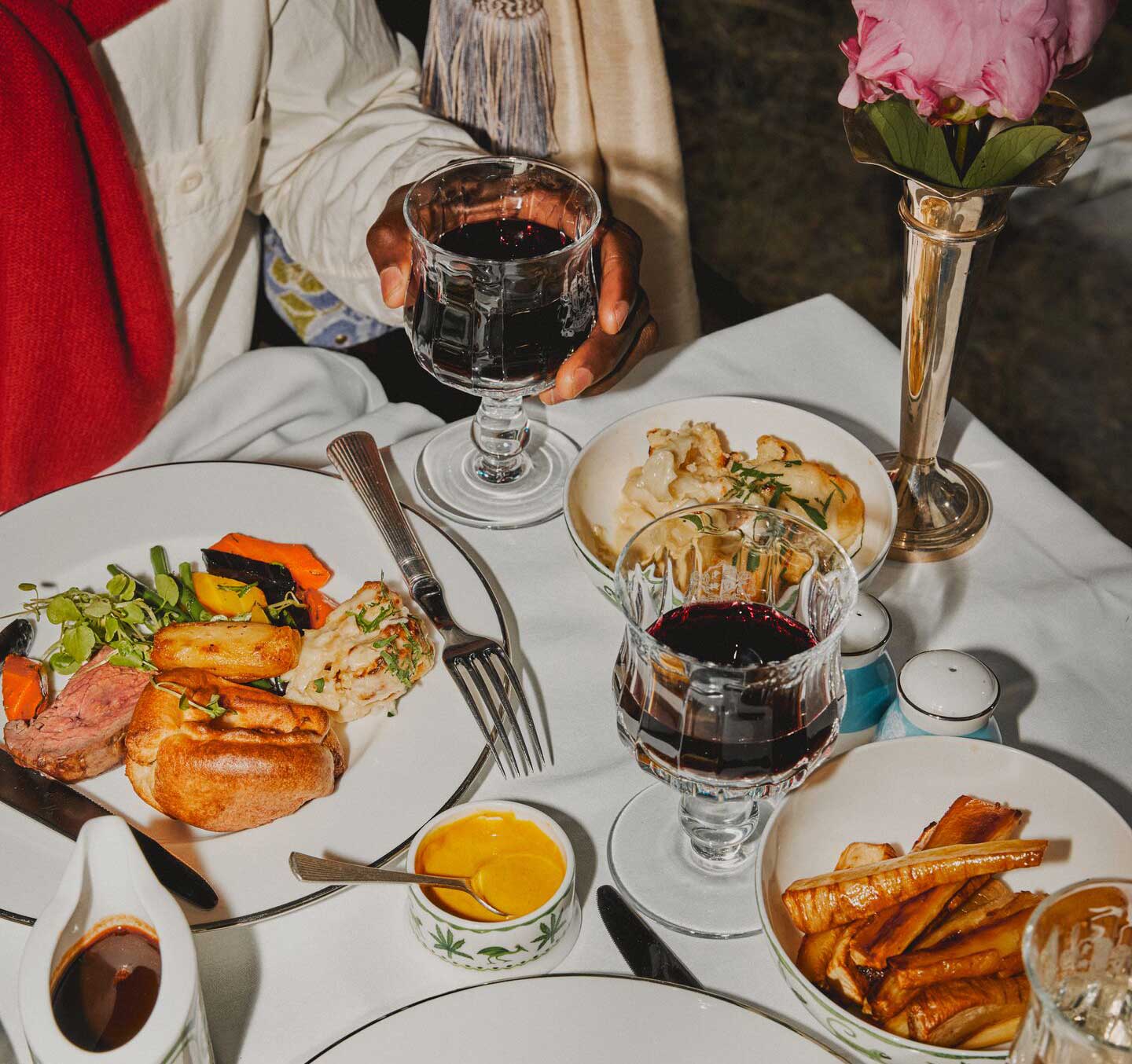 A hand holding a glass of wine over a spread of Sunday roast.