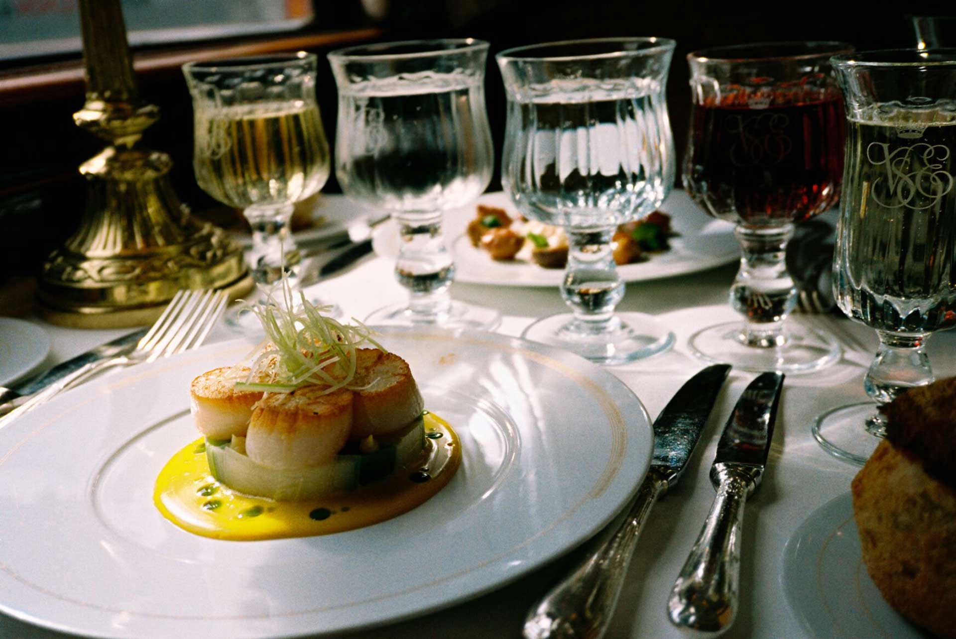 A delicate plate os scallops on a table, cluttered with drinks, plates, and cutlery.