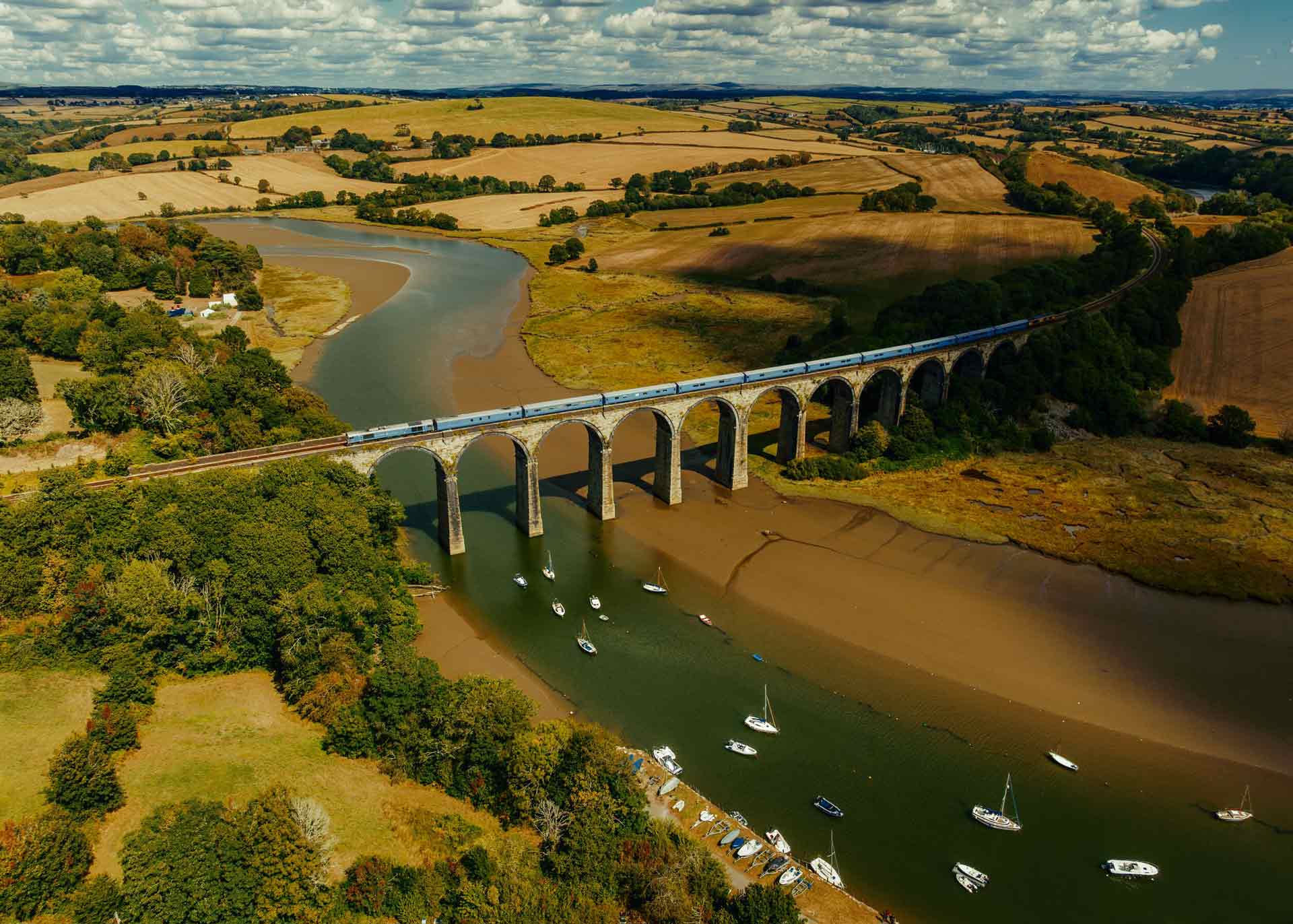 Landscape shot over a river and countryside. A train is crossing a railway bridge over the river and boats sail the river.