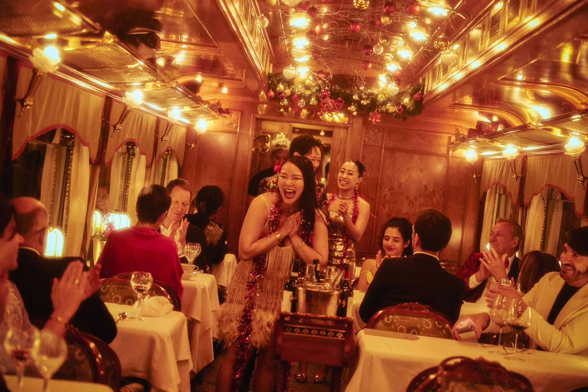 People smiling, laughing, and clapping in a warmly lit Eastern and Oriental Express restaurant carriage.