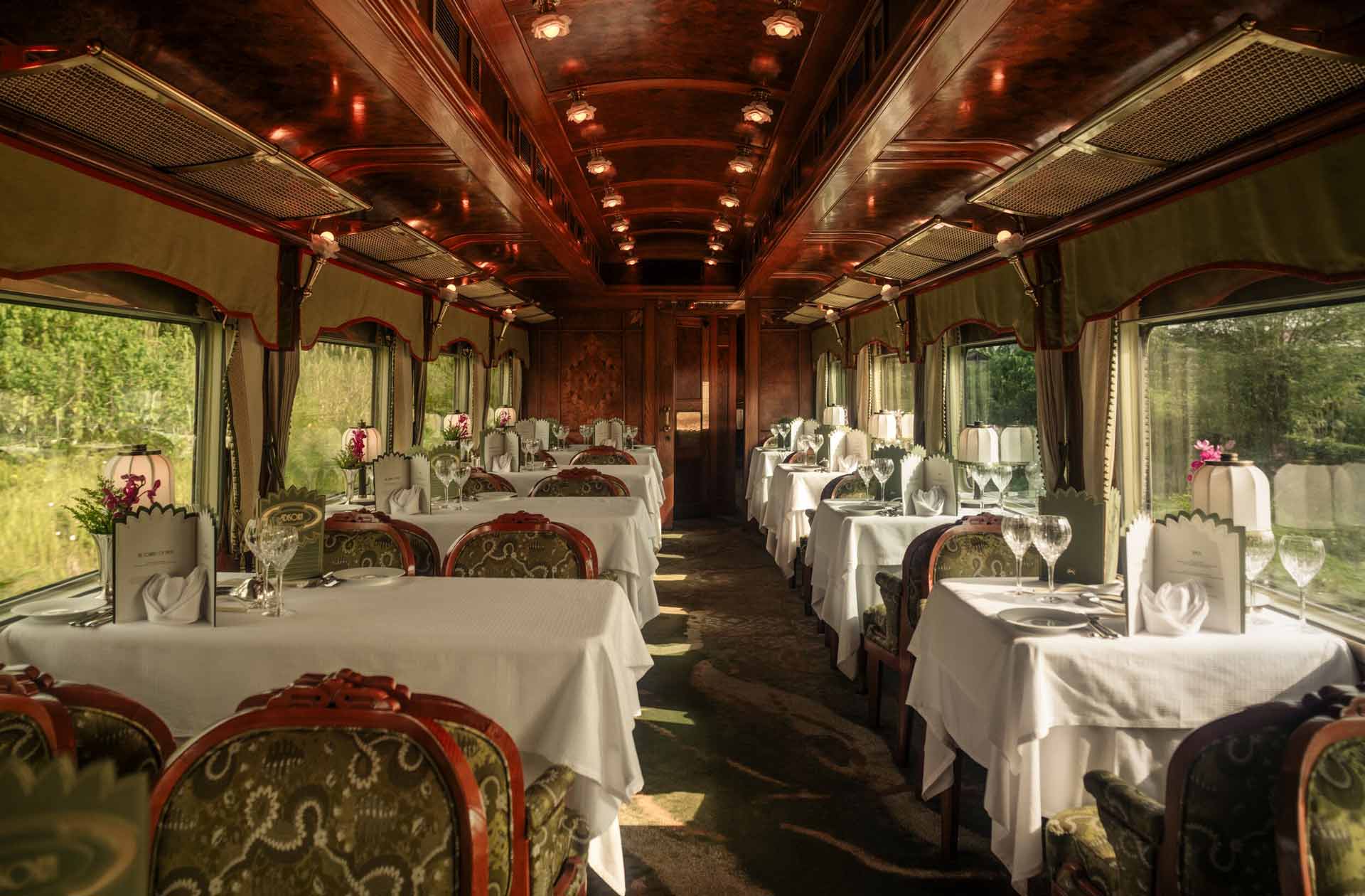 An elegant dining carriage on the Eastern and Oriental Express. Green caprets and upholstery, white tablecloths, and warm lamplight per table.