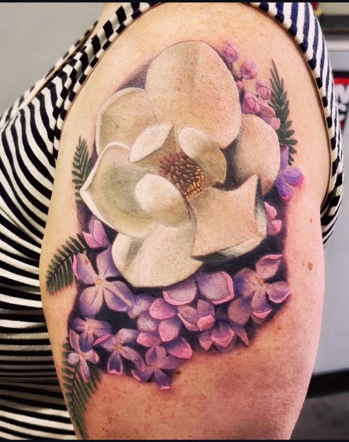 Tattoo of a large white magnolia flower surrounded by clusters of smaller purple flowers and green fern leaves on an upper arm.