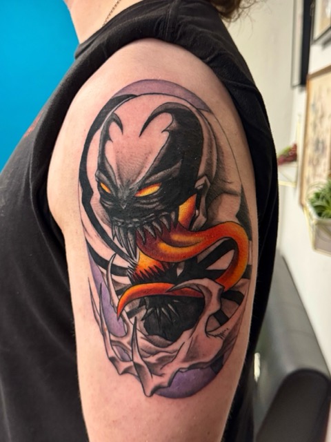 Colorful tattoo on upper arm showing a stylized creature with sharp teeth, glowing yellow eyes, and an extended orange tongue coiling out of its mouth, surrounded by dynamic black and purple shading.