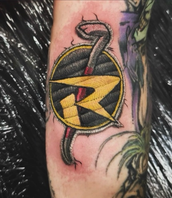 Tattoo of the Robin logo from DC Comics, featuring a stylized yellow R within a black circle with a yellow border, overlaid on a red staff or rod, on a person's forearm with surrounding tattoo designs.