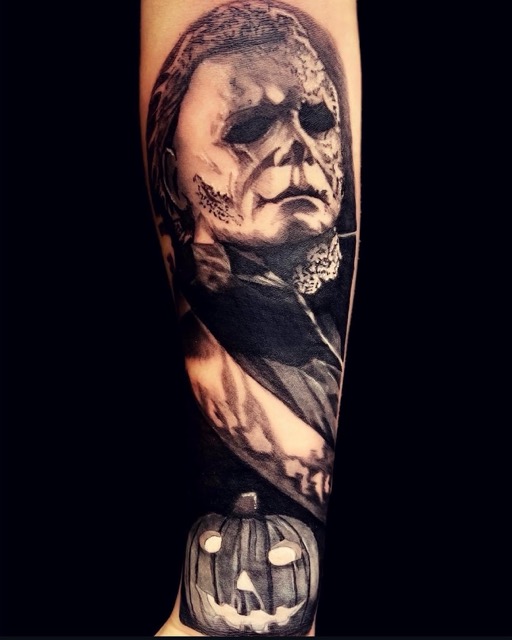 Black and gray tattoo on forearm depicting a detailed portrait of a menacing masked figure with hollow eyes and cracked skin, reminiscent of a horror character, above a glowing, carved jack-o'-lantern with a sinister smile.