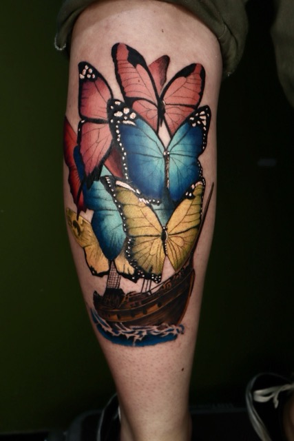 Colorful tattoo on leg depicting a wooden ship sailing on blue waters with sails formed by large butterflies in pink, blue, yellow, and red hues.