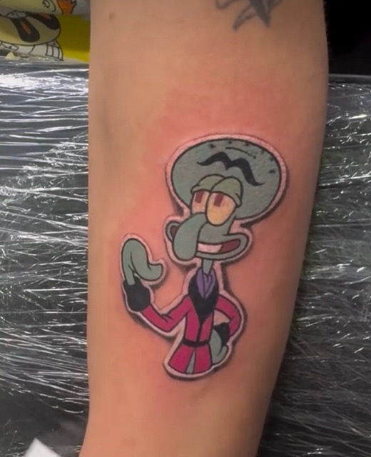 Tattoo on forearm of Squidward Tentacles from SpongeBob SquarePants wearing a red suit and smirking with one hand raised.