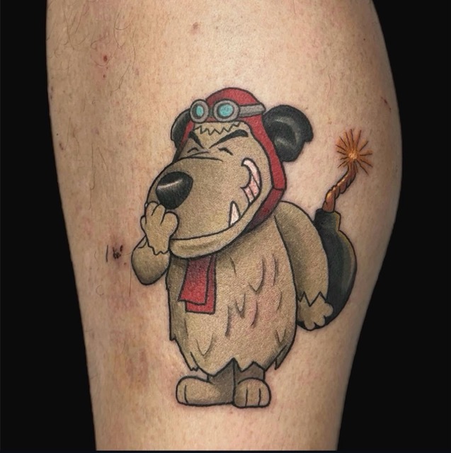 Tattoo of a cartoon dog wearing aviator goggles and a red scarf, holding a lit bomb behind its back, with a mischievous grin on its face.