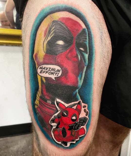 Colorful tattoo on a person's thigh depicting Deadpool's masked face with a speech bubble saying 'MAXIMUM EFFORT!' and a small chibi-style Deadpool holding a gun below it, all set against a blue oval background.