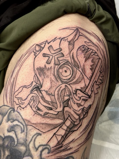 Black ink tattoo on upper thigh featuring a stylized demon or ogre face with exaggerated round eye, sharp teeth, and tribal markings, holding a long blade, surrounded by circular lines.