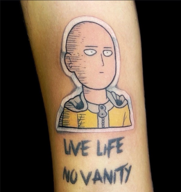 Tattoo of a cartoon character with a bald head and yellow outfit above the phrase "LIVE LIFE NO VANITY" inked on a person's arm.