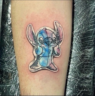 Tattoo of Stitch, the blue alien character from Disney's Lilo & Stitch, with large ears and black eyes on light skin.