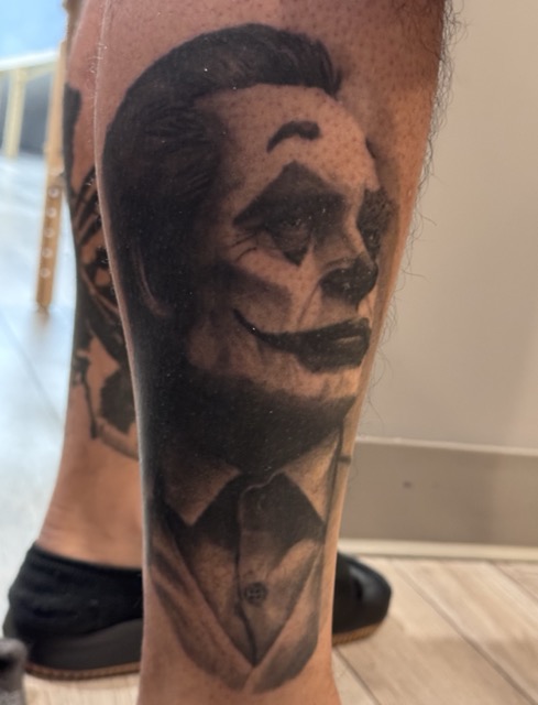 Tattoo of a man's face painted like a skull with dark eye sockets, nose, and mouth, wearing a suit and tie on a person's calf.