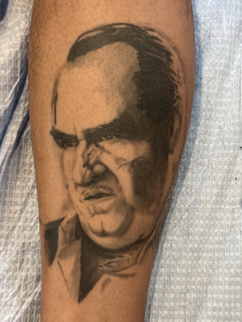 Black and gray portrait tattoo on forearm showing a man's serious face with slicked-back hair and intense expression.