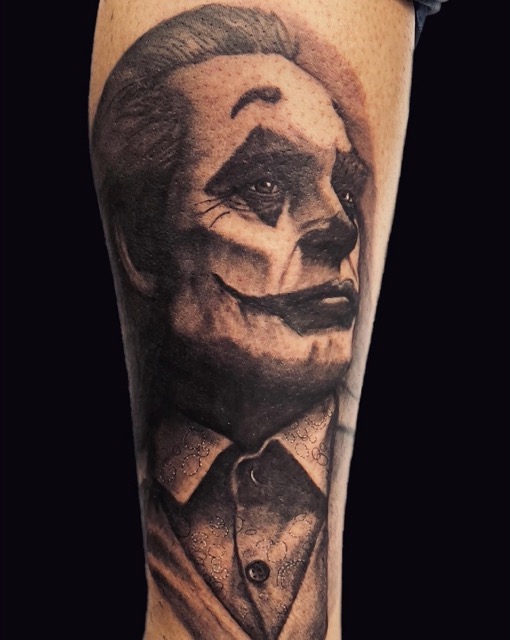 Black and gray tattoo of a person with clown makeup looking upward, wearing a textured shirt with a buttoned collar.