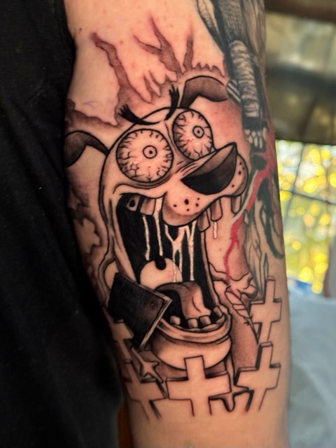 Black and gray tattoo of a cartoon-style dog with wide, bloodshot eyes and an open mouth showing teeth and dripping saliva, surrounded by cross-shaped tombstones on a person's arm.