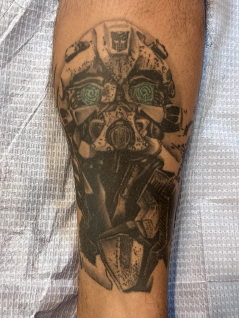 Black and grey tattoo on a person's forearm depicting a stylized robotic or mechanical face with intricate details and greenish eyes.