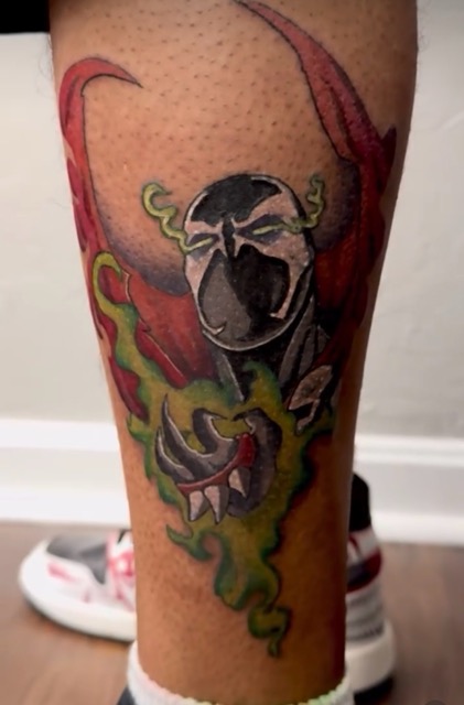 Colorful tattoo on a person's lower leg featuring a stylized black and white mask-like face with green flame-like elements and red shapes extending upward.