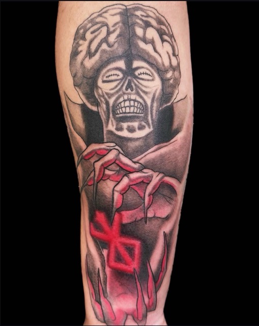 Tattoo on a forearm depicting a skull with closed eyes and a large brain, holding a glowing red symbol between skeletal fingers, with black and red shading.