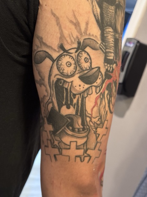Tattoo of a cartoon-style dog with bulging eyes, prominent front teeth, a collar, and a frantic expression surrounded by cross shapes on a person's arm.