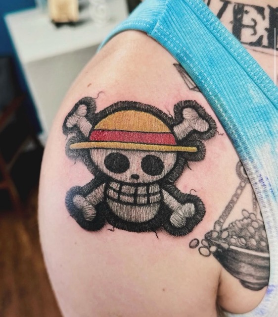 Tattoo on upper arm of a cartoonish skull with large eye sockets and a wide grin, wearing a straw hat with a red band, framed by two crossed bones behind it.