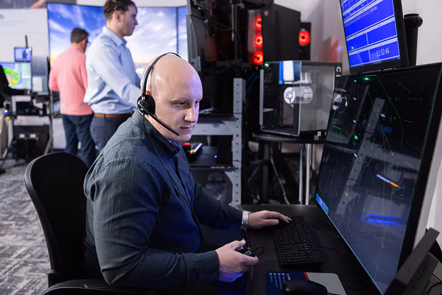 Man wearing a headset training on Adacel's MaxSim air traffic control simulator with a supervisor and a radar trainee also working in the background.