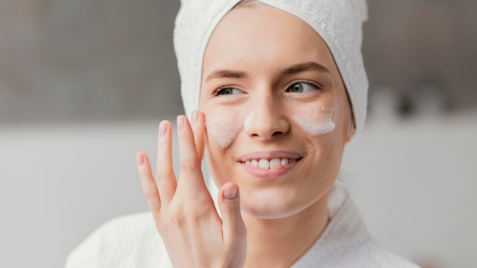 A woman is smiling while applying cleanser to her face as part of a pore-care routine.