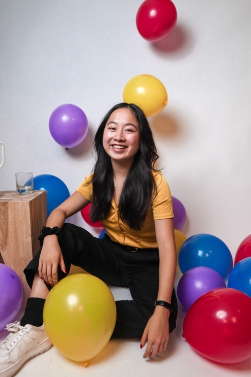 A female with black hair and yellow collared shirt and black pants. Purple, blue, red, and yellow balloons are floating around her.