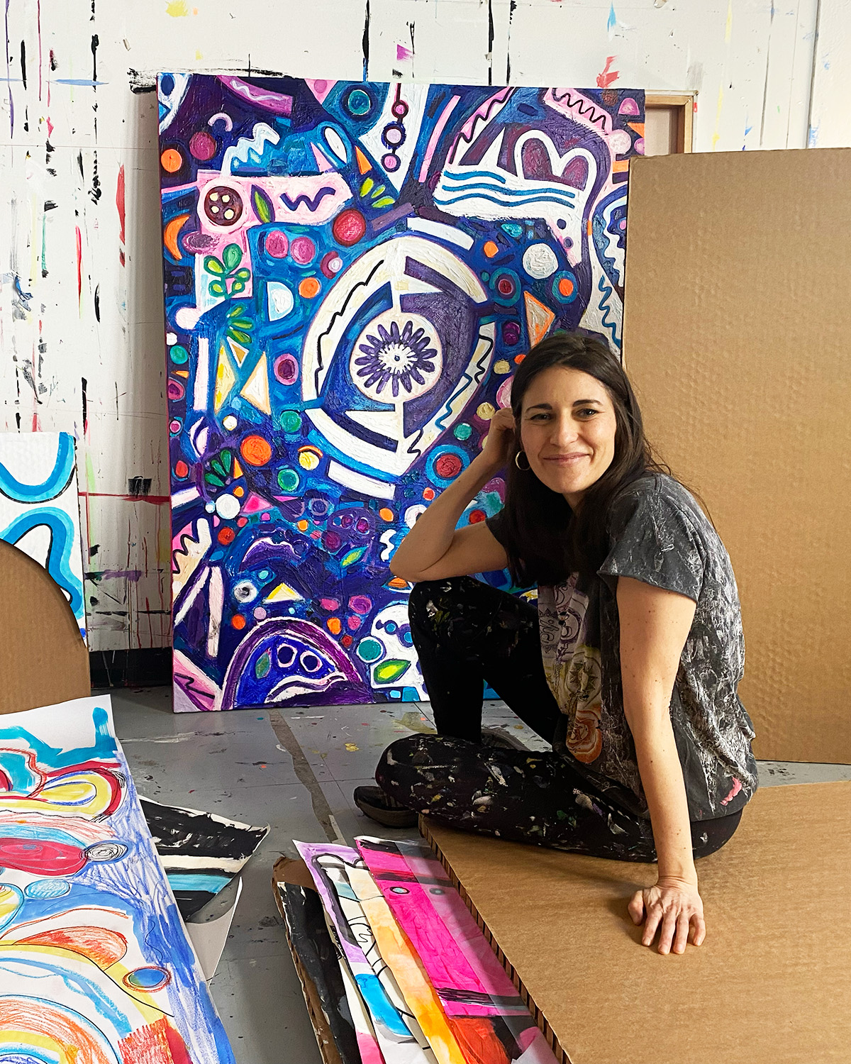Artist Sarah Zeffiro poses sat on the floor of her studio, a large purple and blue illustrative painting sits behind her.