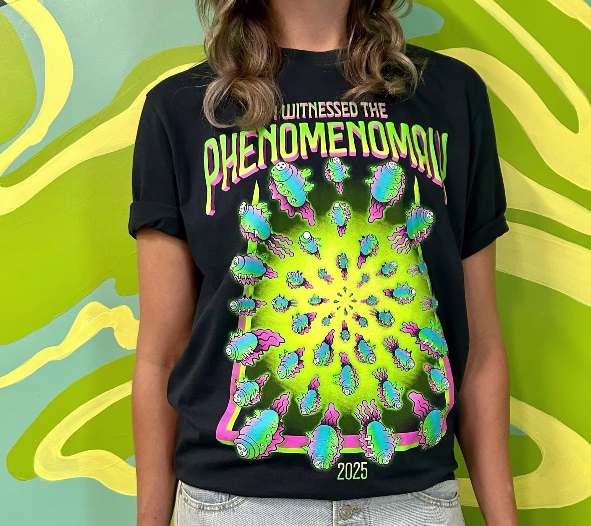 dark t-shirt with the words "I witness the phenomenomaly" against a green and yellow swirled background