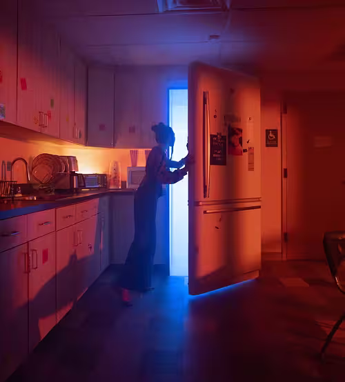 Person opening fridge door in dim kitchen lit by orange and blue light.