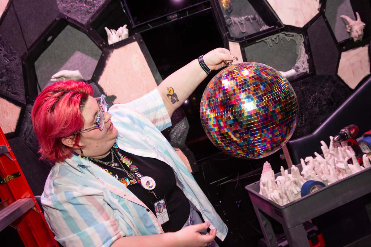 A person with red hair holds up a rainbow disco ball in a room with gray and white tiled walls.