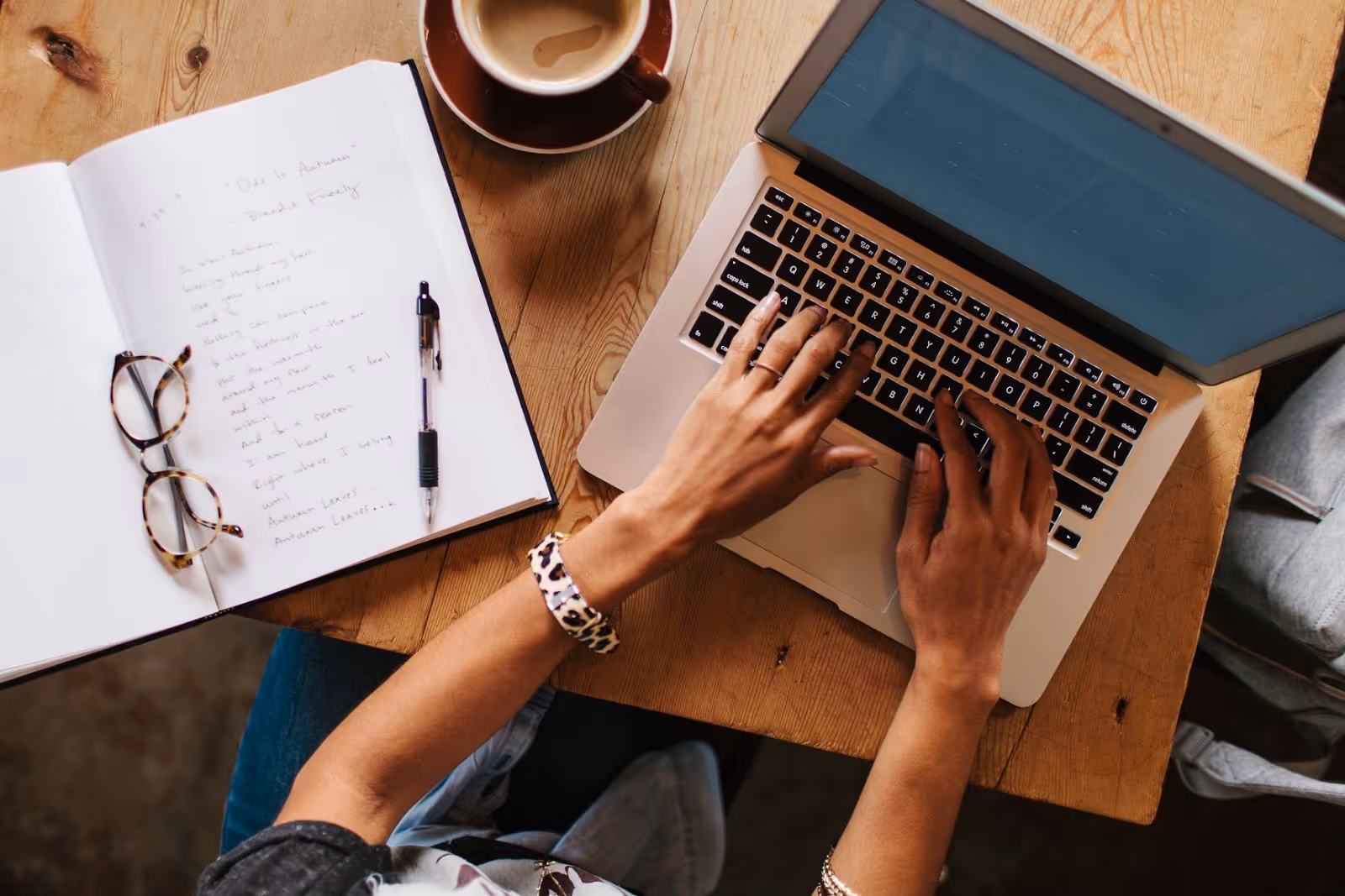 A woman typing on a computer with a cup of coffee and a notebook beside her. 
