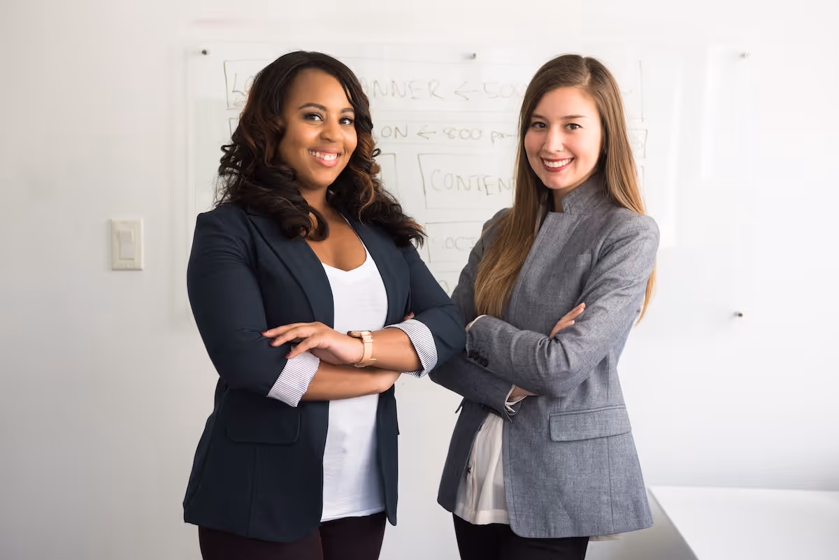 Two women in suits standing in front of a white board, smiling.